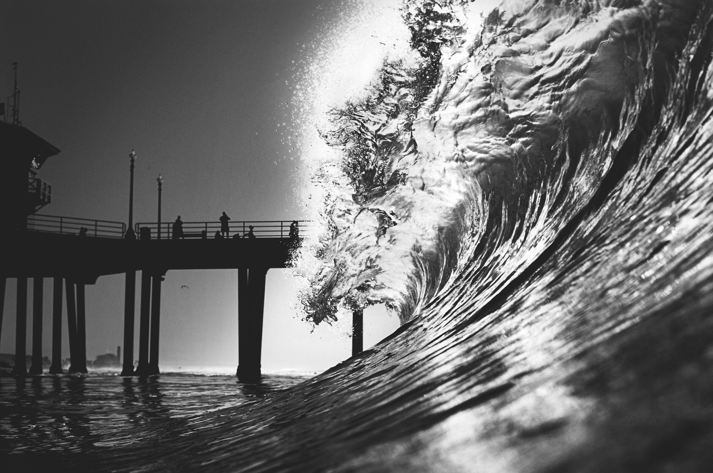 Grande vague déferlant au premier plan, photographie noir et blanc, avec une jetée et des silhouettes en arrière-plan