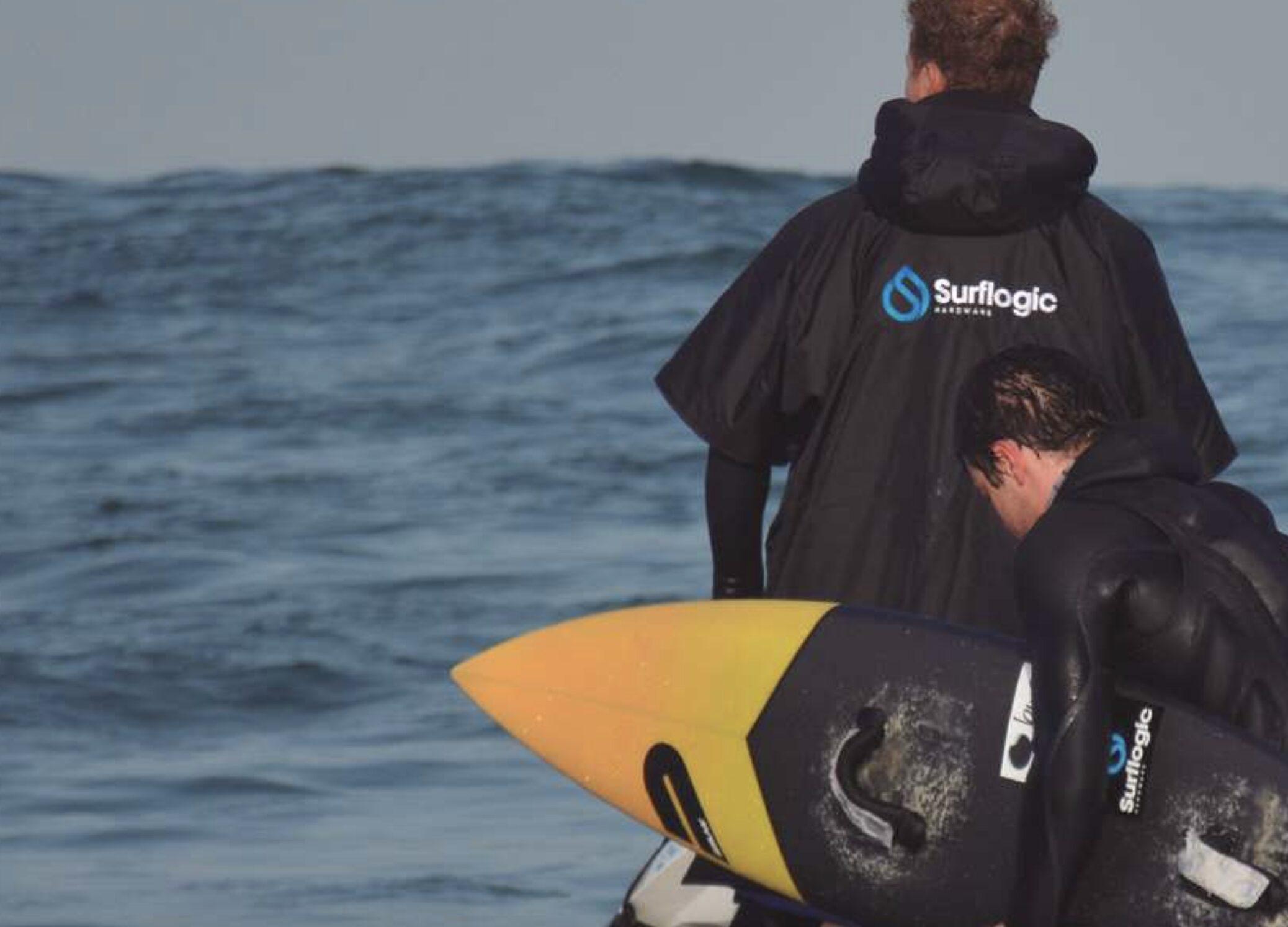 Deux surfeurs en combinaison noire et poncho à capuche, vus de dos au bord de l’eau